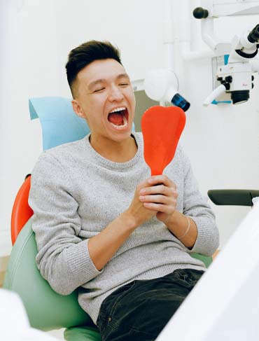 man sitting in dental chair holding mirror looking at mouth
